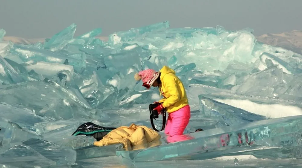 A person in a yellow jacket and pink pants exploring the unique blue ice formations on Lake Baikal during a winter tour.