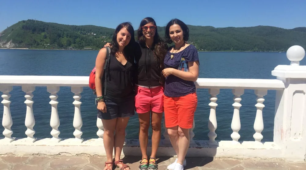 Three women tourists smiling and posing by the waterfront in Listvyanka with Lake Baikal and mountains in the background