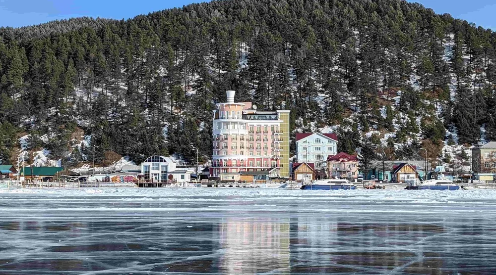 View of Listvyanka village buildings and houses on the shore of frozen Lake Baikal with forested mountains in the background