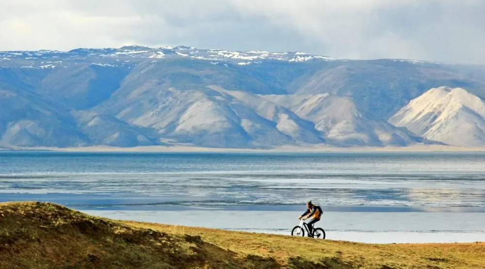 Person riding a bicycle on a scenic coastal path with Lake Baikal and mountains in the background during an Olkhon Island tour